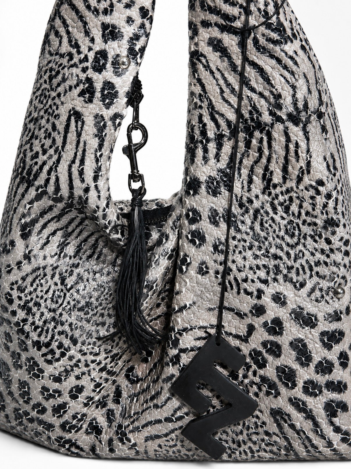 Close up of a slouchy shoulder bag with a black and gray animal print pattern, featuring a black tassel charm and a black elnze logo charm.